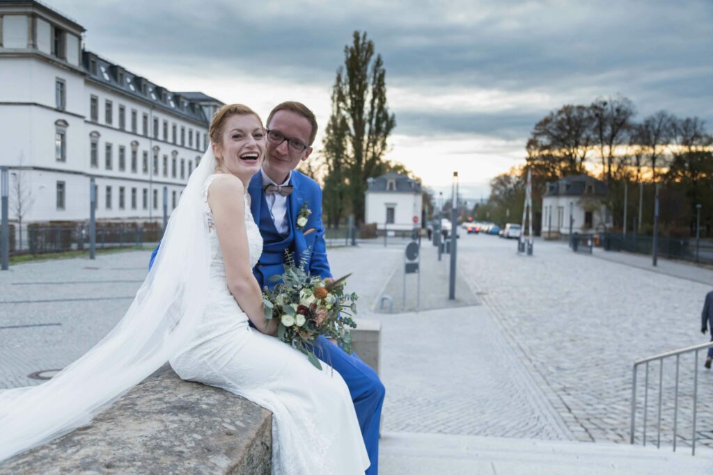 Jan Windisch, Fotograf und Hochzeitsfotograf aus Dresden, Prina, Görlitz, Großenhain und Leipzig, hat hier ein Paar fotografiert. Beide sitzen auf einer Steinmauer und schauen in die Kamera, sie hält noch einen Blumenstrauß in der Hand.