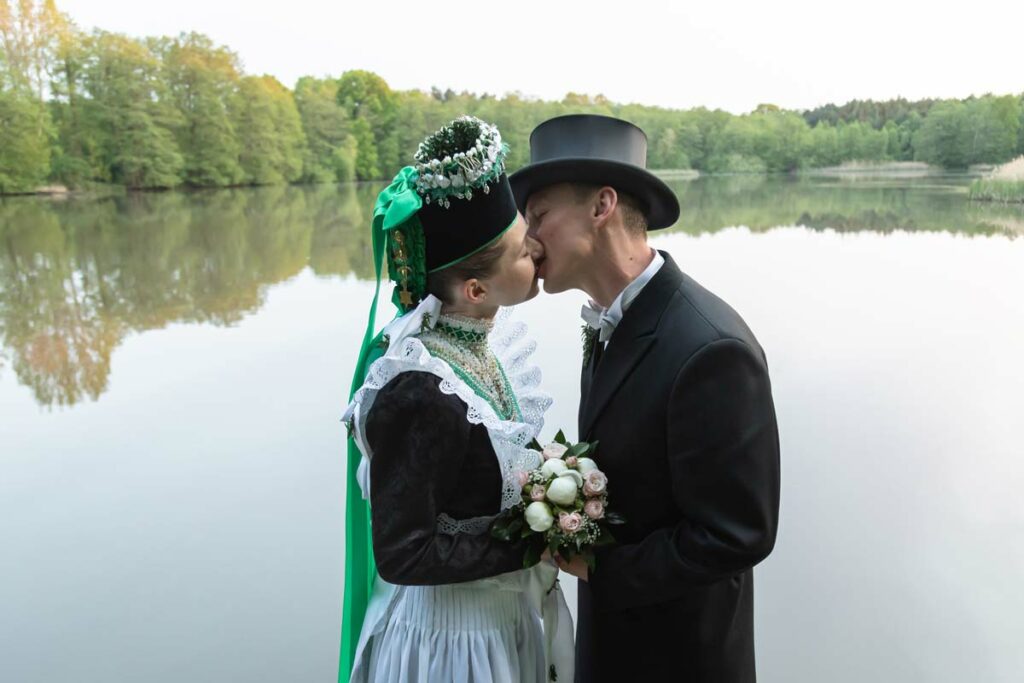 Jan Windisch, Fotograf und Hochzeitsfotograf aus Dresden, Prina, Görlitz, Großenhain und Leipzig, hat hier ein sorbisches Brautpaar fotografiert. Beide stehen gegenüber voneinander und im Hintergrund ist ein See. Das Paar küsst sich.