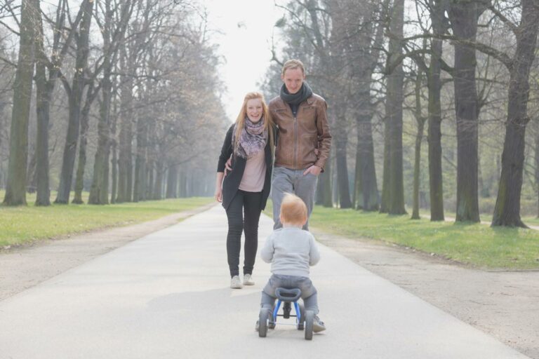 ein kleiner junge fährt mit seinem dreirad und die eltern spazieren einen weg entlang in einem park in dresden - natürliche familienfotos, die familie denkt nicht an den fotografen und haben spaß mit einander