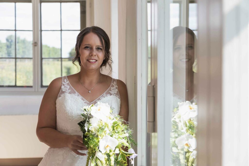 eine Braut steht am Fenster mit ihrem Brautstrauß und schaut in die Kamera