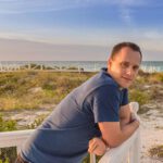 Hochzeitsfotograf Jan Windisch lehnt lässig an einem Holzgeländer und schaut in die Kamera. Im Hintergrund sieht man das Meer von Kuba.