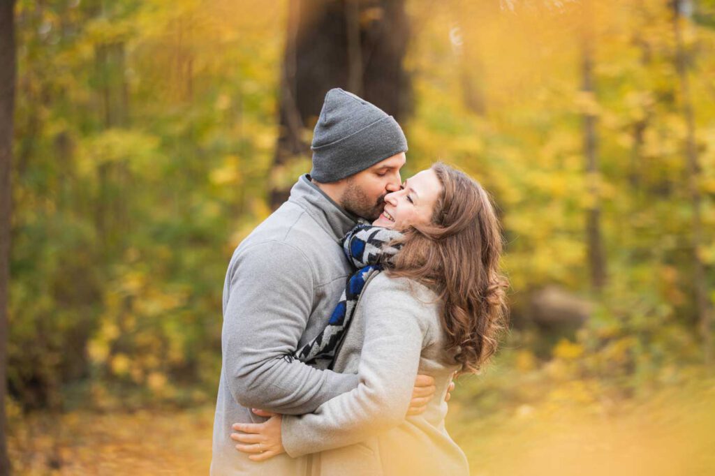 Mann küsst seine Frau und sie stehen eng beieinander - im Hintergrund sieht man eine schöne Herbstfärbung - Fotoshooting im Herbst